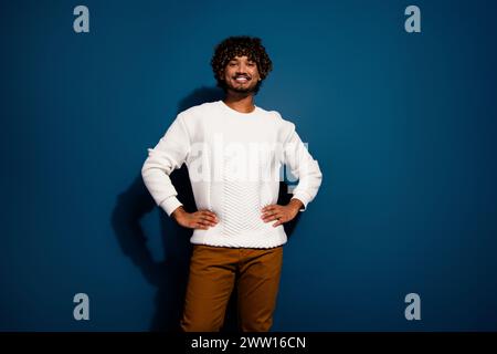 Photo of young confident guy smiling wearing white pullover touching waist posing satisfied isolated on dark blue color background Stock Photo