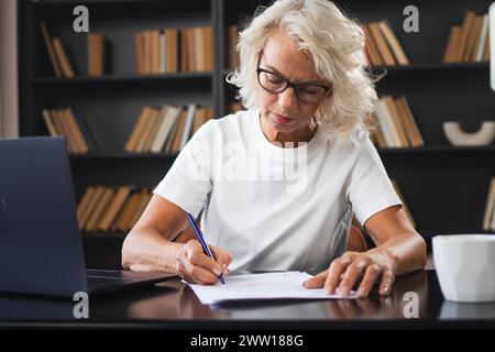 Middle aged senior woman using laptop computer writing notes at workplace. Focused mature old businesswoman work at office making written records doing online work. Mature boss professional worker Stock Photo