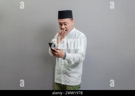 Happy asian muslim man using smart phone cellphone for calls, social media, mobile application online, wearing a koko shirt and peci with shades of th Stock Photo