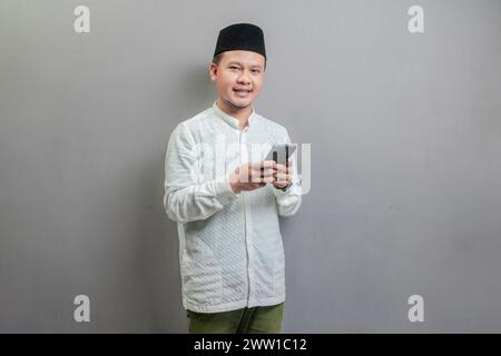 Happy asian muslim man using smart phone cellphone for calls, social media, mobile application online, wearing a koko shirt and peci with shades of th Stock Photo