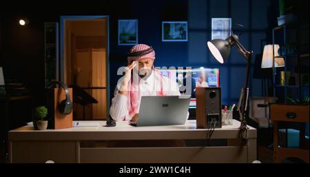 Tired arab businessman suffering migraine working at home office desk. Exhausted Middle Eastern employee in living room answering emails getting headache from overworking Stock Photo