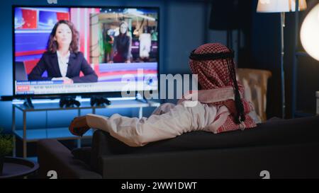 Arab man lounged on cozy sofa, watching news broadcast while relaxing at home after hard day at work. Muslim person chilling on couch, looking at TV channel showing current events Stock Photo