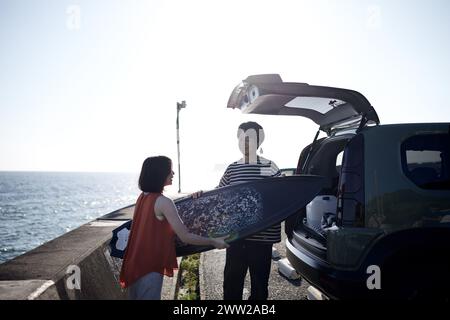 A man and a woman standing next to a car with a surfboard Stock Photo