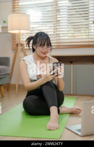 A beautiful, fit young Asian woman in sportswear is chatting with her friends on her smartphone while sitting on a yoga mat, exercising at home. Stock Photo
