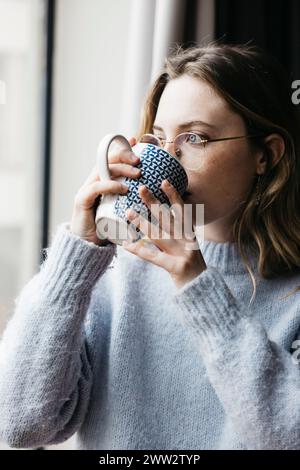 Thoughtful young beautiful blonde woman holding a cup of tea by a window inside their home, creating an intimate and cozy atmosphere Stock Photo