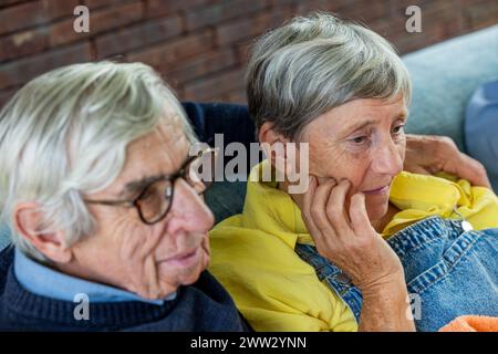 Close-up of a contemplative senior woman with her hand on her cheek, sitting closely next to her male companion. They are both in a thoughtful state, suggesting a moment of reflection or concern, with a cozy indoor backdrop accentuated by a brick wall. Pensive Senior Woman with Companion in Thoughtful Moment. High quality photo Stock Photo