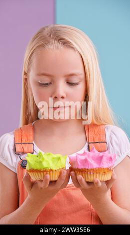 Studio, girl and decision with cupcakes, desert and choice for snack and childhood. Child, sweets and yummy selection for tasty, eating and choosing Stock Photo
