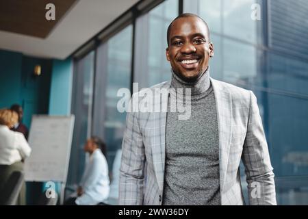 Young African male entrepreneur stands confidently in a modern office boardroom, surrounded by a diverse and professional team. Smiling at the camera, Stock Photo