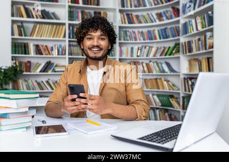 A cheerful young man with curly hair smiles while studying from home, surrounded by books, a laptop, and a smartphone. Stock Photo