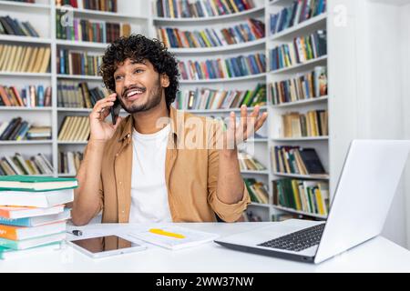 Happy young man with curly hair makes a phone call while working on his laptop in a home office with bookshelves in the background. Stock Photo