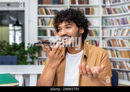Cheerful young man with curly hair speaks into his smartphone using voice command in a library setting. Stock Photo