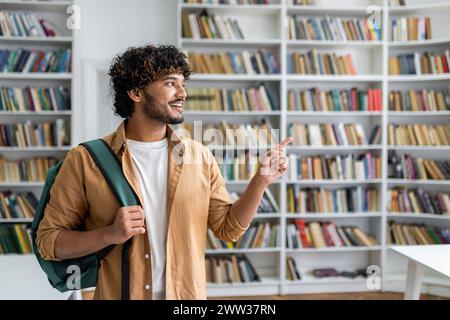 A young adult in a casual outfit with a backpack pointing at something in a library full of books. Stock Photo