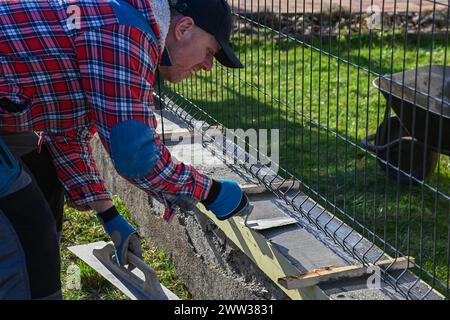 A man in overalls and gloves is repairing the fence in front of the family house. He uses a plane, a trowel and a wheelbarrow.. Stock Photo