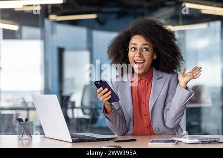Happy African American businesswoman using smartphone, showing surprise and joy at her desk in a contemporary office setting. Stock Photo