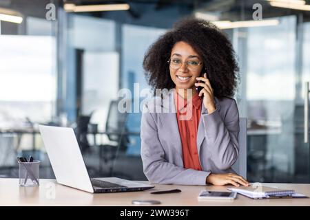 A young professional woman engages in a phone conversation at her office desk surrounded by technology and modern office decor. Stock Photo