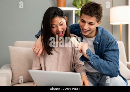 Young carefree diverse couple shopping online with laptop in a sofa. Happy man and woman happy for paying with credit card using a computer at home. Stock Photo