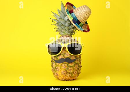 Pineapple with Mexican sombrero hat, sunglasses and fake mustache on yellow background Stock Photo