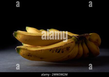 bunch of fresh bananas, ripe tropical fruit with bright yellow peel and deliciously sweet flavor, on dark textured surface with isolated on black Stock Photo