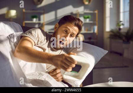 Emotional shocked overslept man holding alarm clock waking up too late lying in bed at home. Stock Photo