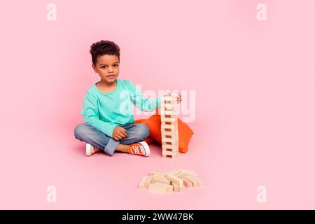 Full size photo of charming small boy play jenga game playroom wooden tower dressed stylish cyan outfit isolated on pink color background Stock Photo