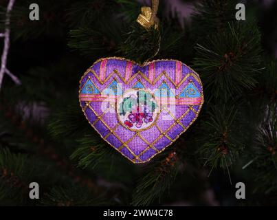 A cross stitched Christmas Timeless Elegance ornament on to a Christmas tree. This heart shape purple Christmas ornament with grapes embroidered and m Stock Photo