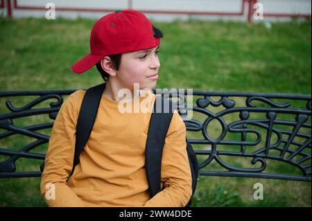 Portrait of a happy teenage schoolboy in a red cap, sitting on a park bench, smiling and looking dreamily to the side. Adorable adolescent multi ethni Stock Photo