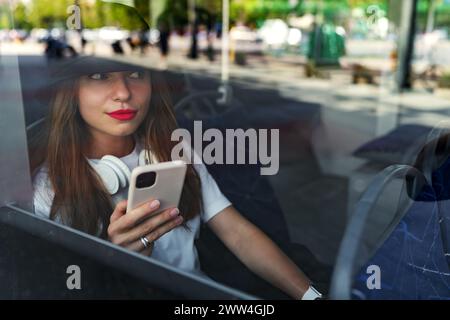 Young pretty urban woman on the bus sits next to the window and uses her smartphone. Photo through the window glass. Stock Photo