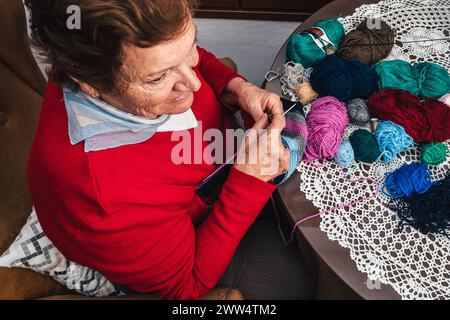 horizontal photo of senior woman knitting with wool and needles and looking to the side sitting in the living room at home Stock Photo