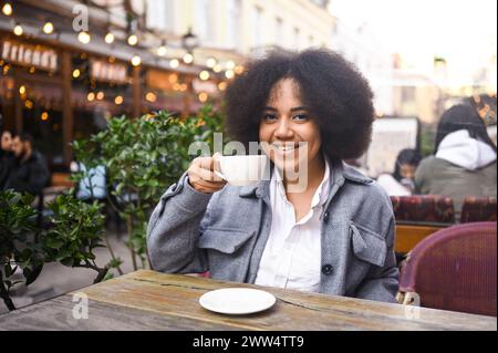 Fashion street style portrait of attractive young natural beauty African American woman with afro hair in tweed jacket posing outdoors in sidewalk cafe. Happy lady with coffee cup in big city Stock Photo