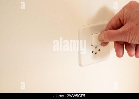 Hand switching on the light on a combined power outlet and switch device, mounted on a white wall. Stock Photo