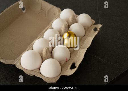 Brown open egg box on a dark table with ten white eggs in it. One egg in the middle is a golden easter egg. Stock Photo