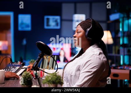 Side view portrait of african american female talk show host speaking with her online audience during a live podcast recording from her home studio, she wears wireless headphones. Stock Photo