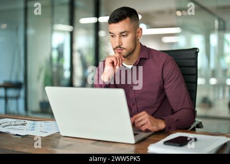 Busy Latin business man thinking working on laptop sitting at office desk. Stock Photo