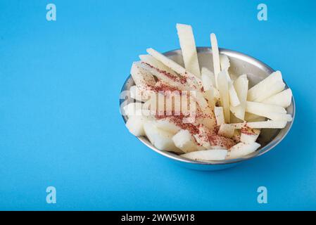 Sliced yam bean with chile and lime in a metal bowl on a blue background. Mexican snack. Jicama. Stock Photo