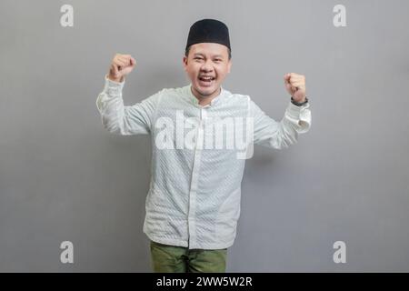 Happy asian muslim man fun successful raised hands show muscles wearing a koko shirt and peci with shades of the fasting month, isolated on a gray bac Stock Photo