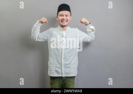 Happy asian muslim man fun successful raised hands show muscles wearing a koko shirt and peci with shades of the fasting month, isolated on a gray bac Stock Photo