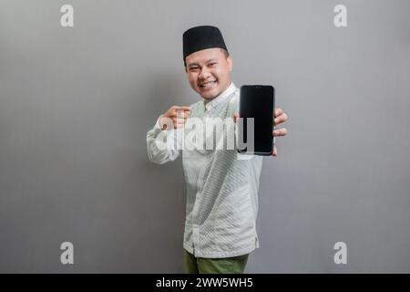Portrait of an Asian Muslim man wearing a koko shirt and peci with shades of the fasting month, smiling with pointing finger to his smartphone, isolat Stock Photo