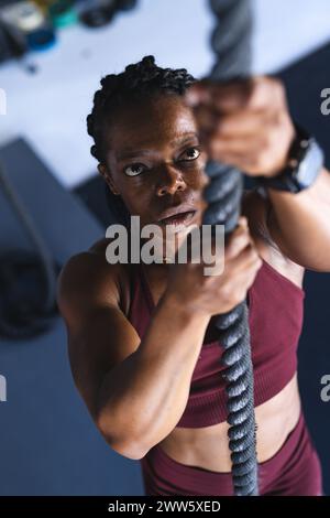 A middle-aged African American strong fit woman is climbing a thick rope in the gym Stock Photo