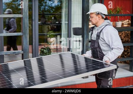 Technicians building solar panel system. Men workers in helmets carrying photovoltaic solar module outdoors. Concept of alternative and renewable energy. Stock Photo