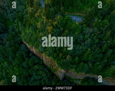 A top-down view of a solitary car on a curving road on top of large rock cliff, surrounded by the dense, vibrant foliage of a forest. Stock Photo