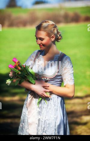 A young woman with blonde hair sits smiling in a dirndl on a wooden jetty on the lakeshore, holding tulips in her arms. Stock Photo