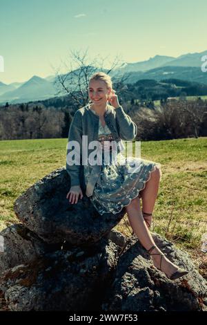 A woman with blonde hair and a dirndl sits on a stone in front of snow-covered mountains. A cardigan keeps her warm. Stock Photo