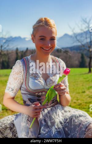 A woman with blonde hair and a dirndl sits on a stone in front of snow-covered mountains in spring. She is holding a tulip in her hand. Stock Photo