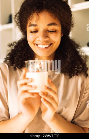 Close-up of charming mixed-race teenager holding candle in glass candlestick and laughing. Handsome African girl stands in stylish bright dressing roo Stock Photo