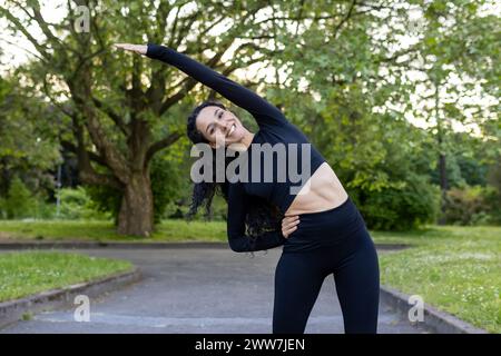 Smiling woman performing a side stretch in a green park setting, showcasing a healthy lifestyle and outdoor fitness routine. Stock Photo