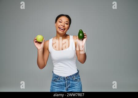 Radiant african american woman with apple and avocado promoting balanced nutrition on grey backdrop Stock Photo