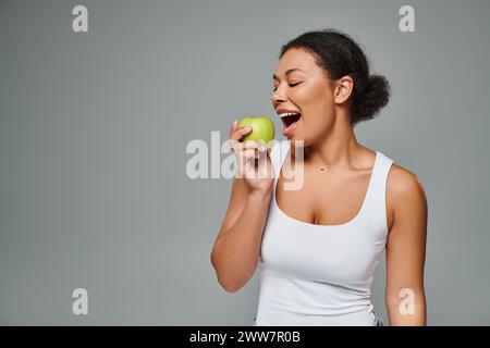 happy african american woman with white teeth biting green apple on grey background, healthy eating Stock Photo