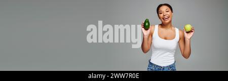 Radiant african american woman with apple and avocado promoting balanced nutrition on grey, banner Stock Photo