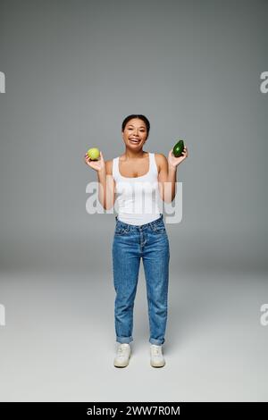 happy african american woman with apple and avocado promoting balanced nutrition on grey backdrop Stock Photo
