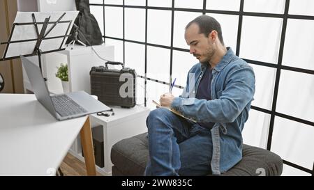 A bald hispanic man with a beard writes notes indoors, reflecting on his work by a laptop and music stand. Stock Photo